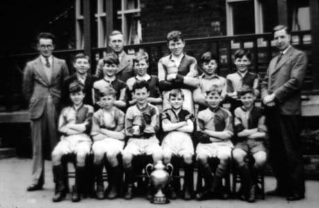 Photograph showing twelve boys, aged approximately ten years, posed in front of a building with large windows; eleven of the boys are wearing football strip; in front of the group is a trophy cup; the boys are accompanied by three men identified as Mr. Potts, Mr. Coverdale, Mr. Peart; the boys have been identified as members of Thornley Junior School