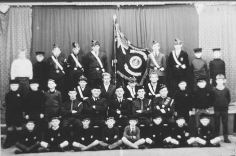 Photograph of a group of twenty seven boys posed against a curtain on a stage in a hall; in the middle of the group are five men, the middle one of whom has been identified as Mr. Speckman; a flag can be seen in the middle of the group; the boys have been identified as members of the Boys' Brigade at their Annual Display at Thornley