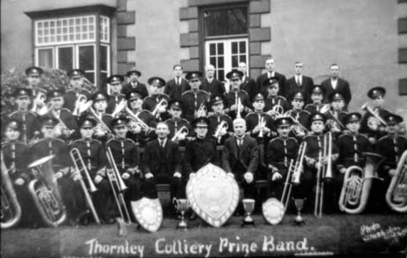 Photograph showing a group of twenty five men in uniform accompanied by nine men in suits posed in front of a house with a bay window; the men in uniform are holding musical instruments and two trophy cups and three shields are in front of the group; the words Thornley Colliery Prize Band and  Photo Chisholm are written on the photograph