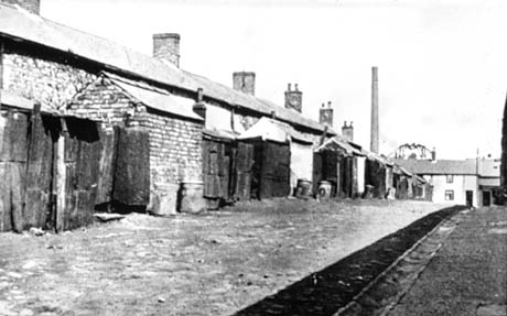 Photograph showing an unmetalled back road running away from the camera and the backs of terraced houses, backs of their outbuildings and decrepit fences; in the distance the facades of houses and a tall chimney can be seen; the photograph has been described as Slum Clearance, Thornley