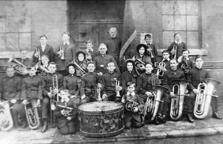 Photograph showing a group of twenty six men, women and children holding musical instruments, posed against the door, wall and windows of a building; an elderly man standing in the doorway of the building is holding a flag, the details of which cannot be seen; the group has ben identified as The Salvation Army Band in Thornley