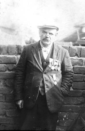 Photograph of a middle-aged man standing against a brick wall; he is wearing a cap, waistcoat and jacket on which five military medals are pinned; he has been identified as Mrs. Smith's Father With 1914-1918 Medals in Thornley