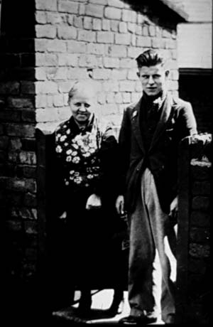 Photograph showing a middle-aged woman and a young man standing side by side in a gateway with a brick wall behind the woman; the young man is wearing a jacket and trousers but the clothing of the woman cannot be discerned; they have been identified as Mrs. Smith's Mother and Brother in Church Street, Thornley