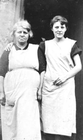 Photograph showing a middle-aged woman on the left of the photograph, wearing a long apron over a dark garment; she has been identified as Mrs. Griffiths; on the right of the picture is a young woman wearing a long pinafore over a dark garment; she has been identified as Emma; they are standing in a doorway of building