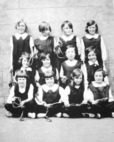 Photograph of twelve girls, aged approximately eleven years, wearing gym slips, standing and sitting in three rows against a wall; they are each holding short, possibly skipping, ropes; they have been identified as taking part in a physical training display at the Council School in Thornley
