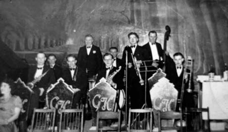 Photograph showing nine men in evening dress with musical instruments sitting and standing on a stage; stage scenery depicting an avenue of trees can be seen behind them; in front of them are four stands with G K on them; a woman can be seen on the left of the picture; the band has been identified as Kitto's Band at Dawdon Welfare