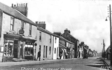 Postcard photograph entitled Thornley, Hartlepool Street. Frith TNY 7, showing on the left of the picture the buildings on the side of a road running away from the camera; a shop nearest the camera is followed by three small terraced houses, which, in turn are followed by two shops; further terraced houses and shops can be seen succeeding each other along the road; none of the names on any of the shops can be read; an indistinct figure is walking near the nearest shop and the back of a motor vehicle can be seen in the distance on the road
