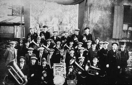 Photograph showing thirty two men wearing suits, ties and caps, and holding musical instruments, standing in front of a colliery banner, the details of which are indistinct, although it appears to have an allegorical figure on it; behind the banner are buildings, possibly colliery buildings; two small children can be seen at the front of the group; the group has been identified as the Colliery Band, Thornley