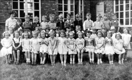 Photograph of thirty five children, aged approximately five years, posed in front of a building with large windows; a woman is standing at the back of the group on the left of the picture; she has been identified as Joan Richardson; the children have been identified as being pupils at school in Thornley