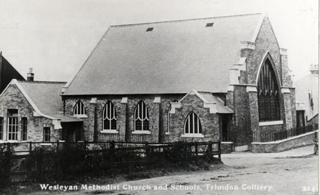 Postcard photograph entitled Wesleyan Methodist Church and Schools, Trimdon Colliery, showing the exterior of the east end and south side of a nineteenth century Gothic church with a low building attached to the west end of the church; a fence and part of a road can be seen in the foreground of the picture