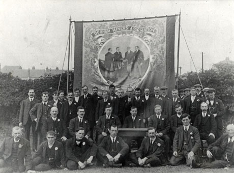 Photograph showing thirty seven men wearing suits, ties and button holes, grouped in front of a banner reading Durham Miners' Association Trimdon Grange Colliery; four men are depicted on the banner; behind the group and the banner are a high hedge and the tops of houses