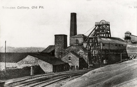 Postcard photograph entitled Trimdon Colliery, Old Pit., showing, in the foreground, the slope of a hill and a railway line curving round to the right of the picture; beyond the railway line, are low buildings of the colliery with the winding gear and a tall chimney behind them; a group of indistinct figures can be seen in front of the low buildings