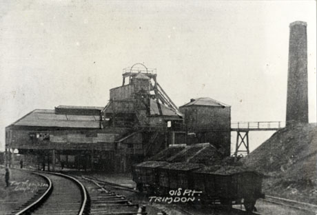 Postcard photograph entitled Old Pit, Trimdon, showing rails running away from the camera with three coal waggons on them; behind the rails are buildings and the winding gear; on the right of the picture is a tall chimney
