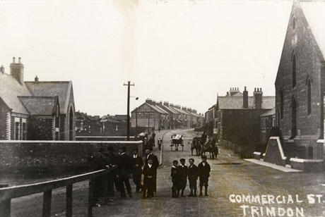 Postcard photograph entitled Commercial Street, Trimdon, showing a road running away from the camera; on the left of the picture is a building, possibly a school, with an open space beyond it; beyond that, where the road climbs are terraces on the end of which the words Clough Clothing can be seen; on the right of the picture is the end of a church and, beyond that, a number of houses set back from the road and the side of the buildings beyond; six men and five boys are standing in the road at the front of the picture and other figures can be seen on the pavements in the distance; three carts can be seen in the road