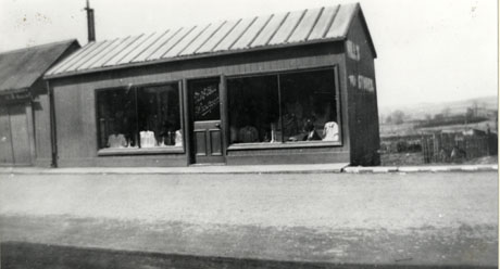 Photograph showing a single-storey wooden building with two large windows at the end of a row of buildings with gardens and open countryside behind it; on the door of the building are the words: M. and C. Hill Showroom; garments can be seen in the windows; the building has been identified as Hill's Drapery Shop, trimdon Colliery