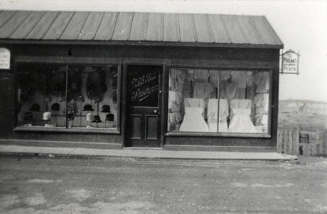 Photograph of the windows of M. and C. Hill Showroom, showing hats on stands in the left window and garments, possibly underwear and nightwear, hanging over rails in the right window; a sign with the name, Pullars, Perth, is on the end of the building; it has been identified as being in Trimdon Colliery