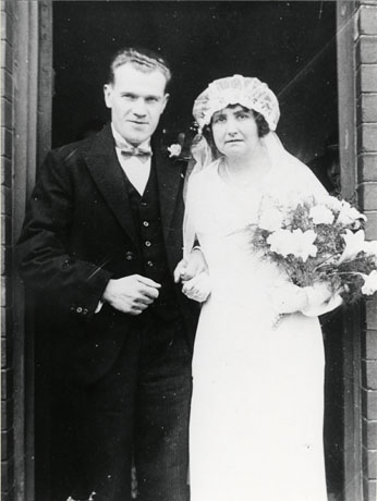 Photograph showing a man in morning dress and a woman in a wedding dress and veil with a bouquet of flowers standing in a doorway; they have been identified as Mr. and Mrs. Hill, Trimdon Colliery, 10 May 1933