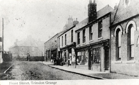 Postcard photograph entitled Front Street, Trimdon Grange, showing a street running away from the camera, showing an indistinct house and the end of the street and, possibly, a level crossing at the end of the street; the facades of five shops and of, possibly, a church can be seen on the right of the picture; the windows of the shops cannot be seen in detail; a group of indistinct people are on the pavement in the distance