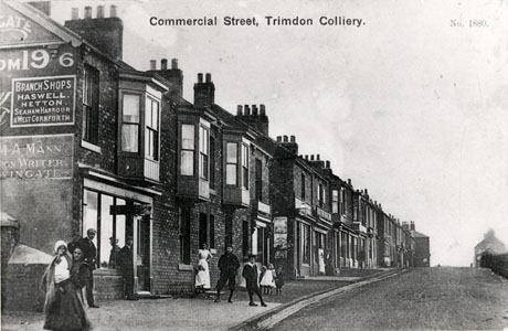 Postcard photograph entitled Commercial Street, Trimdon Colliery. No. 1880, showing the facades of shops on the left of the photograph; the windows of the shops cannot be seen in detail; three men, two women and five children are standing on the pavement near to the camera; two further people can be seen indistinctly in the distance