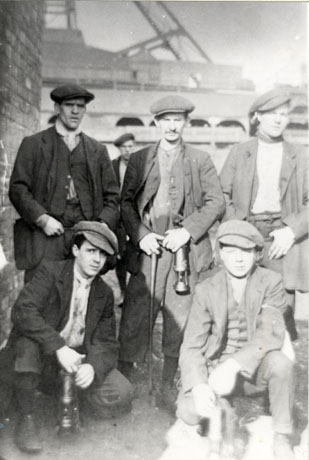 Photograph of three men in work clothes standing with the buildings and winding gear of a colliery behind them; in front of the men, two boys, also in work clothes, are squatting on the floor; all five people are carrying miner's lamps; they are dressed in jackets, waistcoats and caps; they have been identified as miners at Trimdon and as follows: Back Row, Left to Right: Tommy Soulsby; William Harper, Senior; J. Kell; Front Row, Left to Right: Dickie Harper and Billy Harper