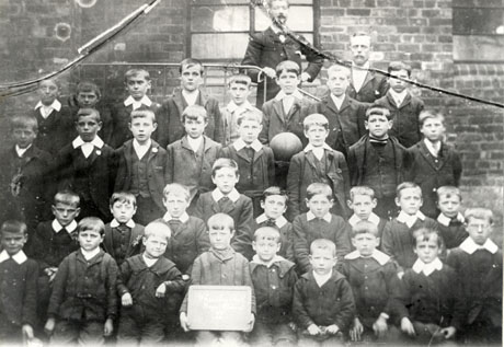 Photograph of thirty three boys, aged approximately ten years, posed in front of the wall of a brick building; two men are standing behind the group; a child on the front row is holding an illegible notice; the children have been identified as pupils in Class 2 in Wheatley Hill Boys' School