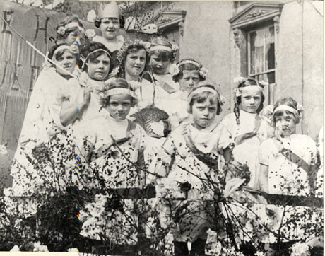 Photograph of eleven girls, aged between approximately seven years and twelve years, standing near the facade of an imposing building; behind the girls is a banner and in front of them is what appears to be a fence with branches attached to it; all the girls are wearing light dresses with headbands in their hair; a child at the back of the group is wearing a paper crown and a girl immediately in front of her is wearing a wreath in her hair and holding a basket; the photograph has been identified as depicting a May Queen in Wheatley Hill