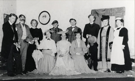 Photograph of six women and five men posed in costume on a stage in front of scenery; they have been identified as the cast of Pygmalion by George Bernard Shaw; the cast are dressed in the clothes of the first decade of the twentieth century; three women in long frocks and large hats are sitting on a sofa centre stage; behind them are the men in suits of the period and a woman dressed as a housekeeper, a woman dressed as a maid and a woman dressed as an upper-class woman considerably older than those on the sofa; they have been identified as members of the drama club in Wheatley Hill
