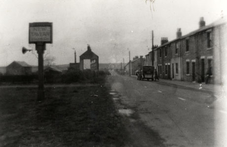 Photograph showing a road running away from the camera with terraced houses on the right of the picture; on the left is an inn sign on a pole reading New Inn; behind the sign two indistinct buildings and the end of a third can be seen; a motor van is parked on the right of the road with its back to the camera