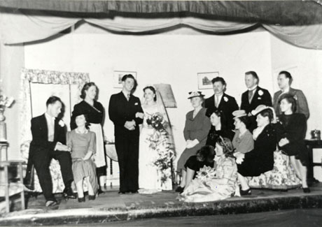 Photograph of the cast of the play Quiet Wedding in costume, posed on stage in front of the scenery; there are a bride and groom in the centre surrounded by four men and seven women in the costume of the nineteen forties, dressed for a wedding