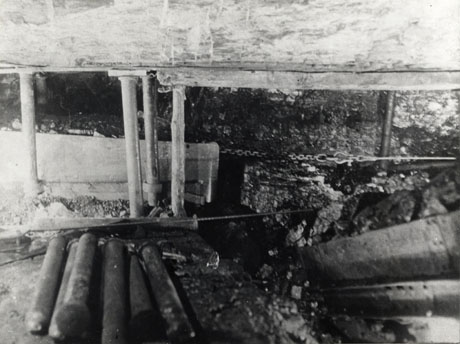 Photograph of the interior of a seam of coal showing pit props, the wall of coal and a chain stretched across the coal; further props can be seen at the front of the picture