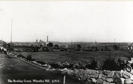 Postcard photograph entitled The Bowling Green, Wheatley Hill. 10564, showing the bowling green surrounded by beds of flowers; behind the green, the chimney, winding gear, and buildings of the colliery can be seen; two indistinct men can be seen standing on the green