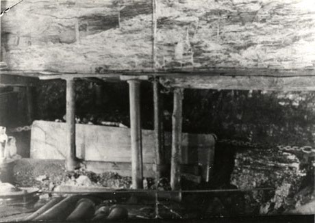 Photograph showing the interior of a coal seam with pit props and a waggon full of coal being pulled by a chain; further props can be seen at the front of the picture