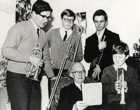 Photograph of a middle-aged man holding a trumpet standing at the left of the picture; next to him are two young men holding a trombone and a trumpet; in front of the two young men are an elderly man, holding a piece of music, and a boy, aged approximately ten years holding a trumpet, both sitting on a sofa; they are in a room on a house and have been described as Kitto Family With Instruments