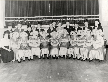 Photograph of a group of ten elderly women seated in a semi-circle, dressed in satin dresses and bonnets; a woman in a blouse and skirt is seated on the left of the semi-circle; behind the women are eleven men seven of whom are wearing top hats; another woman is standing with the men; they are posed in front of a stage with drawn curtains; they have been described as Over 60s Concert Party, Wheatley Hill