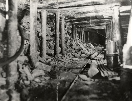 Photograph showing a seam of coal running away from the camera, supported by pit props, and with pieces of coal falling from the walls, across the rails on the ground; the scene has been identified as being at Wheatley Hill Colliery