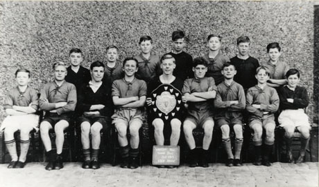 Photograph showing fifteen boys aged approximately between eleven and fourteen years, sitting posed in front of a wall; a boy in the middle of the group is holding a shield and has a notice reading: Wheatley Hill Senior Boys Oct 1949 Eden House