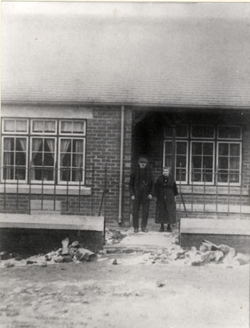 Photograph showing an elderly man and an elderly woman standing in front of a building with two windows, the roof and a wall with railings in view; rubble can be seen in front of the low wall; the photograph has been identified as the Aged Miners' Homes, Wheatley Hill