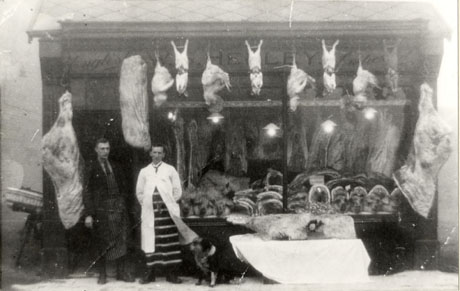 Photograph showing two men wearing striped aprons and overalls standing at the left of the photograph, in front of the window of a butcher's shop; joints and sides of meat are inside the shop window, and sides of meat, chickens and rabbits are hanging outside the shop; above the window, the words, W. S. Hedley English Butcher, are written; a bicycle with a basket on its front can be seen at the side of the shop on the left; the shop has ben identified as being in Front Street, Wheatley Hill