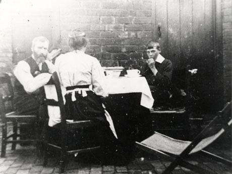 Photograph showing a man in shirt sleeves sitting at the left of the picture on a wooden chair at a table covered with a cloth and laid with crockery; next to him is a woman with her back to the camera wearing a blouse and long skirt, also sitting at the table; on the right of the picture is a boy aged approximately eleven years, sitting at the table and eating; the table has been laid in the back yard of a house; the photograph has been identified as Family in Ford Street, Wheatley Hill