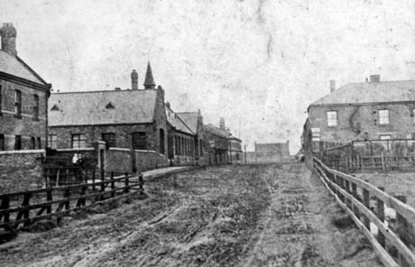 Photograph showing an unmade-up road receding from the camera; on the left are a large house behind a high wall, a large low building, possibly a school, and two other unidentifiable buildings; on the right, are a fence running parallel to the road and a large building beyond the fence; at the end of the road a building, possibly a church, can be seen at right angles to the road; the photograph has ben identified as The Farm, Wheatley Hill