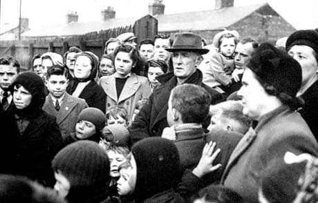 Photograph showing a crowd of men, women and children standing with a fence behind them, beyond which the roofs of terraced houses can be seen; the members of the crowd are wearing overcoats and hats; the photograph has been described as Vesting Day, Wheatley Hill; Vesting Day was 1st January 1947 when the coal industry was nationalised
