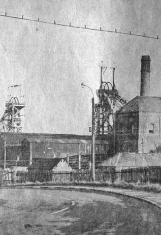 Photograph showing a road curving away from the camera to the left; beyond the road two winding gear, low buildings, a tall chimney and a winding house of a colliery can be seen; the colliery has been identified as Wheatley Hill Colliery