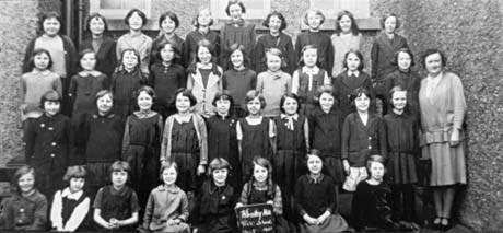 Photograph showing thirty nine girls, aged approximately ten years, posed against the wall of a building; a woman, identified as Miss hope is standing at the right of the picture; a girl on the front row is holding a notice reading: Wheatley Hill Girls' School Standard 5 1931