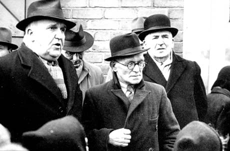 Photograph of six men, wearing overcoats and hats, standing in front of a brick wall, with the backs of the heads of children in front of them;the photograph has been described as Vesting Day, Wheatley Hill; Vesting Day was 1st January 1947 the day the coal industry was nationalised