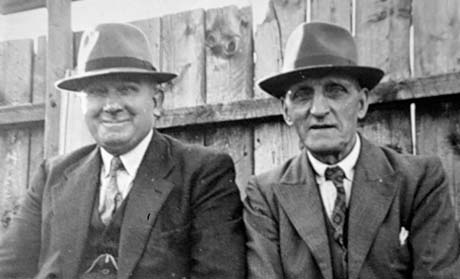 Photograph ot the head and shoulders of two middle-aged men in front of a fence; they are both wearing suits, ties and trilby hats; they have been identified as Joe Fletcher and Mr. Tyson photographed while watching cricket