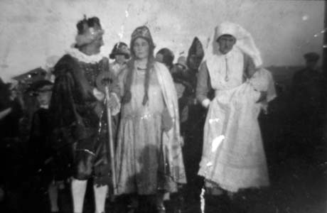 Photograph showing a man wearing knee breeches, a ruff, and a crown, accompanied by a woman wearing long plaits, a cloak and a long dress; on the right of the man and woman, is another woman dressed as a nurse carrying an infant; behind them are indistinct figures, also in fancy dress; they have been identified as taking part in a carnival at Wheatley Hill