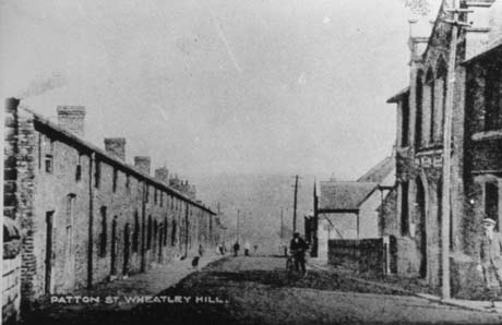 Photograph showing a road running away downhill; on the left is a terrace of houses and on the right a large building, possibly a church and a smaller building further in the distance; the road has been identified as Patton Street, and the church as the Primitive Methodist Church in Wheatley Hill