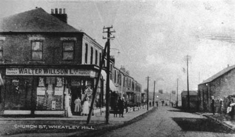 Postcard photograph entitled Church Street, Wheatley Hill, showing a street running away from the camera, showing a Walter Willson shop on the corner of the street on the left; the contents of the windows cannot be seen in detail; two figures are standing in the doorway; beyond the shop is a terrace of houses; on the opposite side of the road is a large barn-like building and an indistinct building beyond that; three indistinct figures can be seen on the pavement