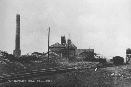 Postcard photograph entitled Wheatley Hill Colliery, showing a large chimney standing alone on the left of the picture; in the centre are buildings with the winding gear of the colliery; at the front of the picture, is a railway track which runs away across the picture to the distance at the right