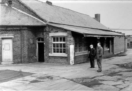 Photograph showing the exterior of the end of a single-storey building showing windows on two sides and a doorway near which a notice can be seen but not read; part of another building can be seen attached to the first near the doorway; two men wearing overalls and caps are standing near the building which has been identified as the Lamp Cabin at Wheatley Hill Colliery